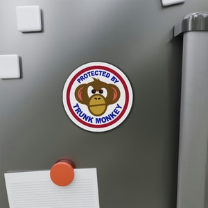 May include: A white round magnet with a red border and blue text. The text reads "Protected by Trunk Monkey". A cartoon monkey face with brown fur, red cheeks, and black eyes is in the center of the magnet.