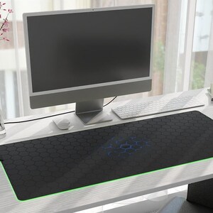 May include: A black gaming mouse pad with a honeycomb pattern and green LED lighting around the edge. The mouse pad is on a white desk with a computer monitor, keyboard, and a cup.