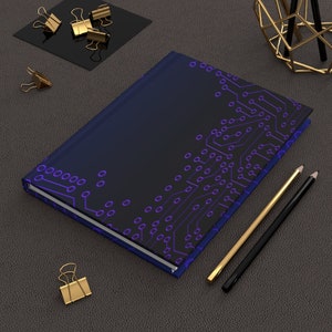 May include: A dark blue notebook with a purple circuit board design. The notebook is lying on a gray surface with two pencils and gold paper clips.