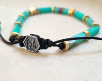 Op de afbeelding: Een turquoise en gouden kralen armband met een zilveren zeshoekige bedel met de letters "MA".
