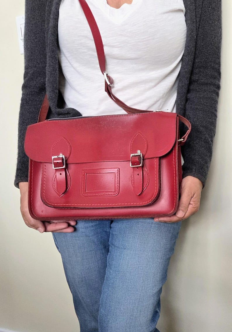 Puede incluir: Un malet&iacute;n de cuero rojo con una solapa frontal con dos cierres de hebilla, un asa superior y una correa de hombro ajustable. El bolso incluye un portaetiquetas rectangular en la solapa frontal y herrajes de metal plateado.
