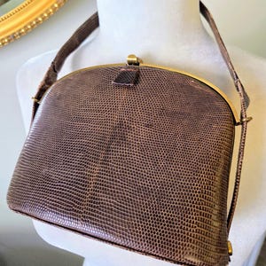 May include: A brown, vintage handbag with a reptile-like texture. The bag features a curved top, a gold-coloured clasp, and a matching shoulder strap. The bag is displayed on a white mannequin.