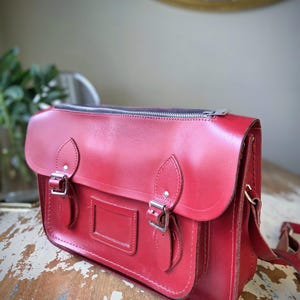 Puede incluir: Un malet&iacute;n de cuero rojo brillante con cierre de cremallera superior, una solapa frontal con dos correas de hebilla ajustables y un tarjetero rectangular. El bolso incluye enganches laterales para una correa de hombro y herrajes plateados.