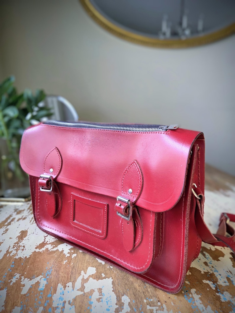 Puede incluir: Un malet&iacute;n de cuero rojo brillante con cierre de cremallera superior, una solapa frontal con dos correas de hebilla ajustables y un tarjetero rectangular. El bolso incluye enganches laterales para una correa de hombro y herrajes plateados.