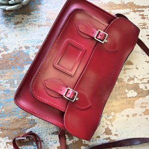 Puede incluir: Un bolso tipo cartera de cuero rojo oscuro con una solapa frontal, dos correas con hebillas ajustables y una ventana rectangular para identificaci&oacute;n. El bolso incluye una correa larga y fina para el hombro y herrajes de metal plateado.
