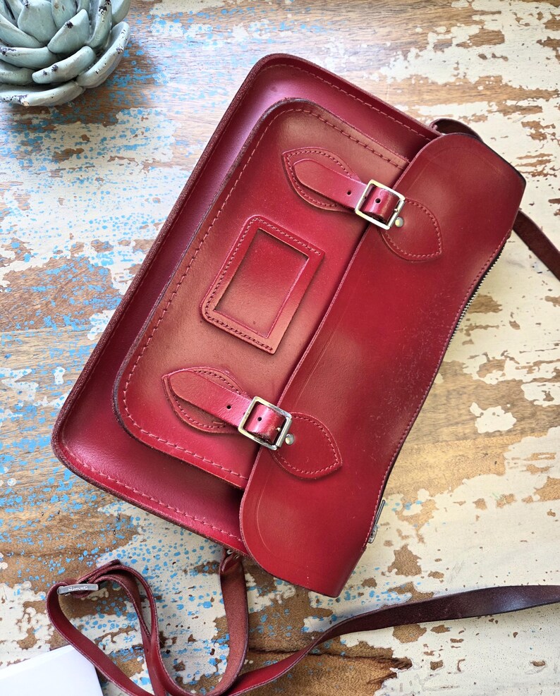 Puede incluir: Un bolso tipo cartera de cuero rojo oscuro con una solapa frontal, dos correas con hebillas ajustables y una ventana rectangular para identificaci&oacute;n. El bolso incluye una correa larga y fina para el hombro y herrajes de metal plateado.