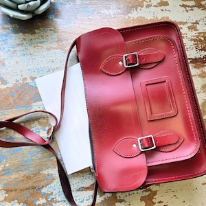 Puede incluir: Un malet&iacute;n de cuero rojo oscuro con una correa larga para el hombro, que presenta una solapa frontal asegurada por dos hebillas de metal plateado y una ventana de identificaci&oacute;n rectangular. El bolso tiene un acabado liso y costuras visibles.