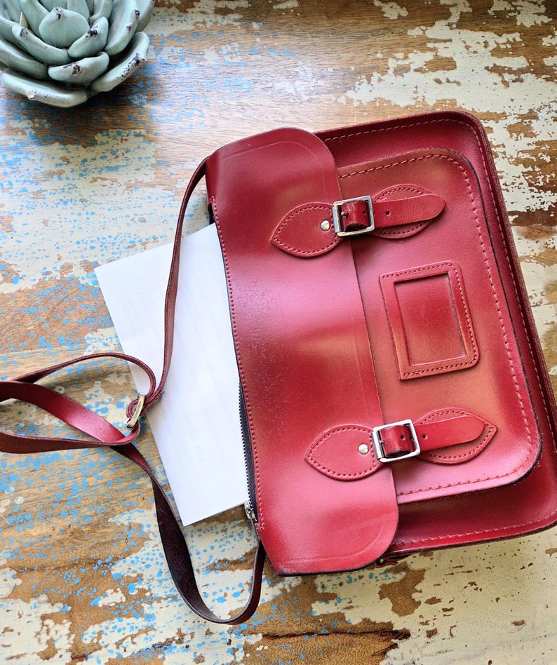 Puede incluir: Un malet&iacute;n de cuero rojo oscuro con una correa larga para el hombro, que presenta una solapa frontal asegurada por dos hebillas de metal plateado y una ventana de identificaci&oacute;n rectangular. El bolso tiene un acabado liso y costuras visibles.