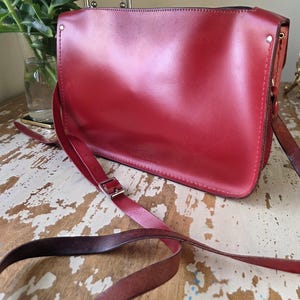 Puede incluir: Un bolso bandolera rectangular de cuero rojo con una solapa frontal lisa, costuras visibles a lo largo de los bordes y una correa larga ajustable con una hebilla de metal plateado. El bolso tiene un dise&ntilde;o estructurado con remaches met&aacute;licos.