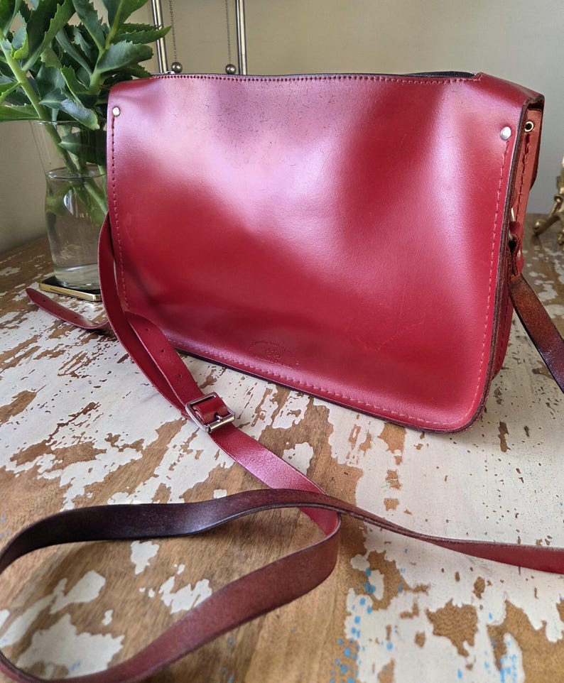 Puede incluir: Un bolso bandolera rectangular de cuero rojo con una solapa frontal lisa, costuras visibles a lo largo de los bordes y una correa larga ajustable con una hebilla de metal plateado. El bolso tiene un dise&ntilde;o estructurado con remaches met&aacute;licos.