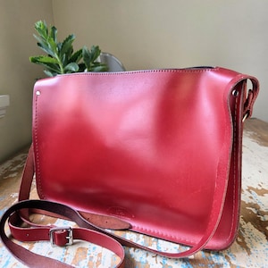 Puede incluir: Un bolso de hombro rectangular de cuero rojo con acabado liso, que cuenta con una correa larga ajustable con una hebilla de metal plateado. El bolso tiene costuras visibles a lo largo de los bordes y un peque&ntilde;o logotipo en el frente.