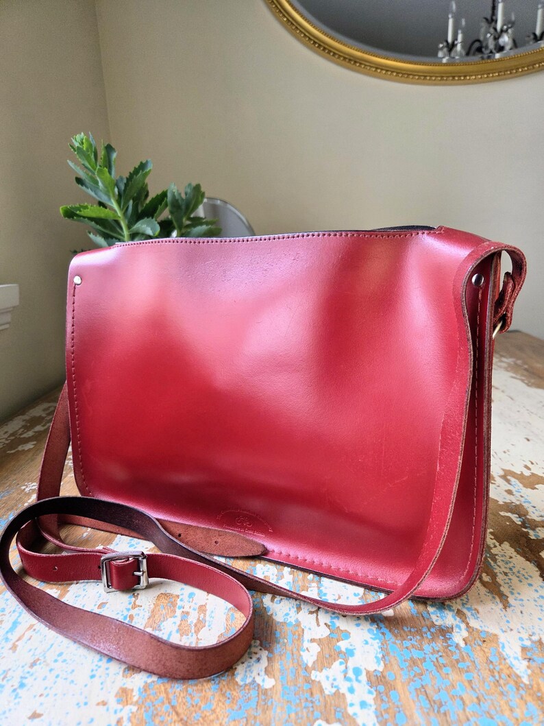 Puede incluir: Un bolso de hombro rectangular de cuero rojo con acabado liso, que cuenta con una correa larga ajustable con una hebilla de metal plateado. El bolso tiene costuras visibles a lo largo de los bordes y un peque&ntilde;o logotipo en el frente.