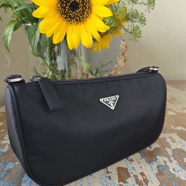 May include: A black nylon handbag with a rectangular silhouette, a top zip closure, and a silver-toned metal Prada logo plaque on the front. The bag features a matching black fabric strap attached with silver hardware at the top corners.