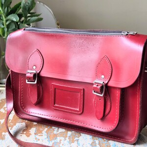 Puede incluir: Un malet&iacute;n estructurado de cuero rojo con cierre de cremallera superior, una solapa frontal con dos correas de hebilla ajustables y un tarjetero rectangular. El bolso incluye una correa larga para el hombro y herrajes de metal plateado.