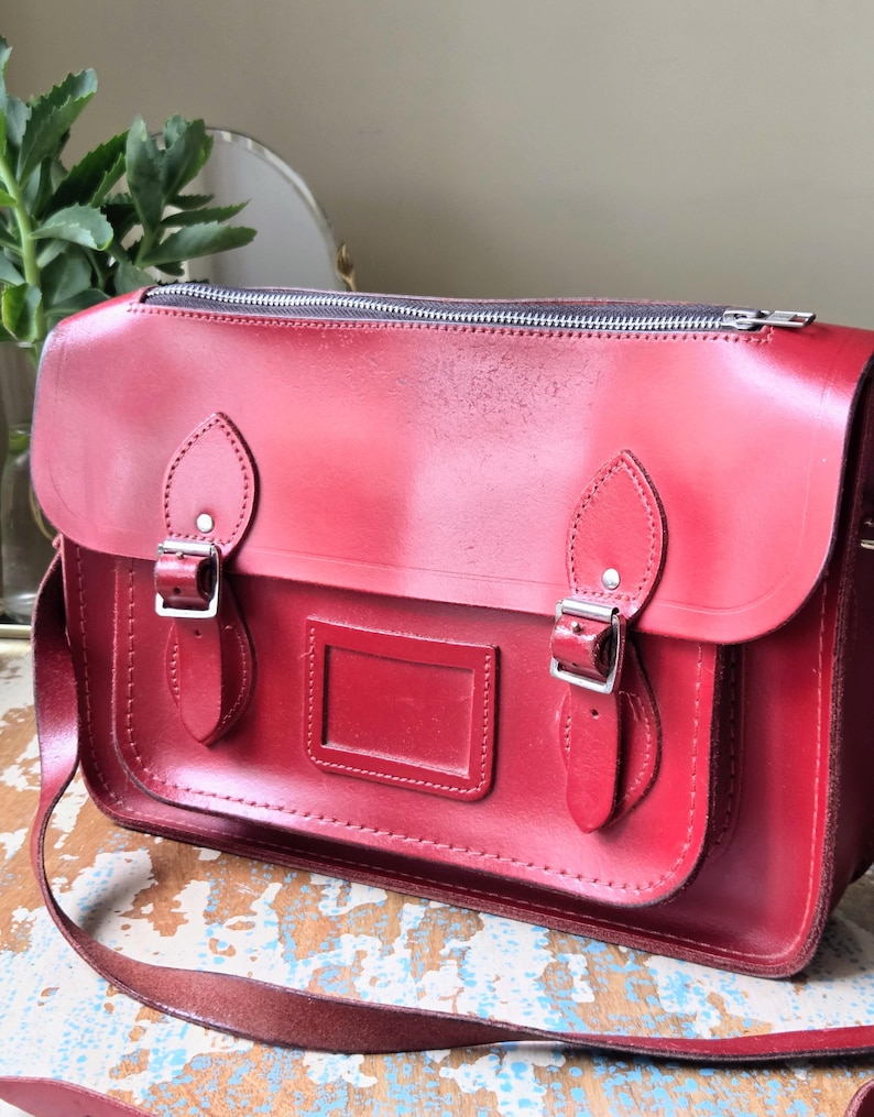 Puede incluir: Un malet&iacute;n estructurado de cuero rojo con cierre de cremallera superior, una solapa frontal con dos correas de hebilla ajustables y un tarjetero rectangular. El bolso incluye una correa larga para el hombro y herrajes de metal plateado.