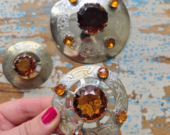 May include: Three vintage brooches with silver-tone metalwork and amber-colored gemstones. The brooches vary in size and design, with intricate details and a central large stone surrounded by smaller gems. One brooch is held by a person.