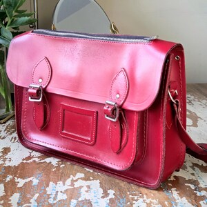 Puede incluir: Un malet&iacute;n de cuero rojo brillante con cierre de cremallera superior, una solapa frontal con dos hebillas met&aacute;licas plateadas ajustables y un tarjetero rectangular centrado en el bolsillo delantero. El bolso tiene costuras visibles.