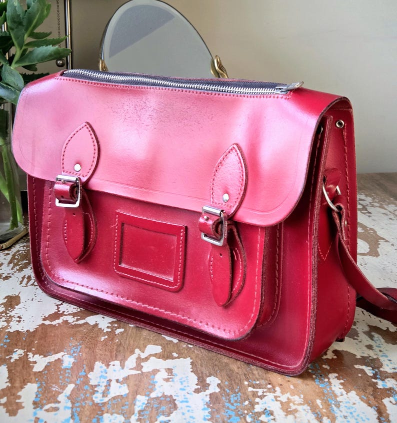Puede incluir: Un malet&iacute;n de cuero rojo brillante con cierre de cremallera superior, una solapa frontal con dos hebillas met&aacute;licas plateadas ajustables y un tarjetero rectangular centrado en el bolsillo delantero. El bolso tiene costuras visibles.