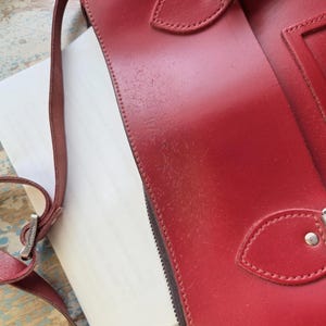 Puede incluir: Un bolso de cuero rojo con cierre de hebilla de metal plateado, una correa de hombro fina y ajustable con un conector de metal plateado grabado con la palabra Cambridge, y costuras visibles a lo largo de los bordes de la solapa.