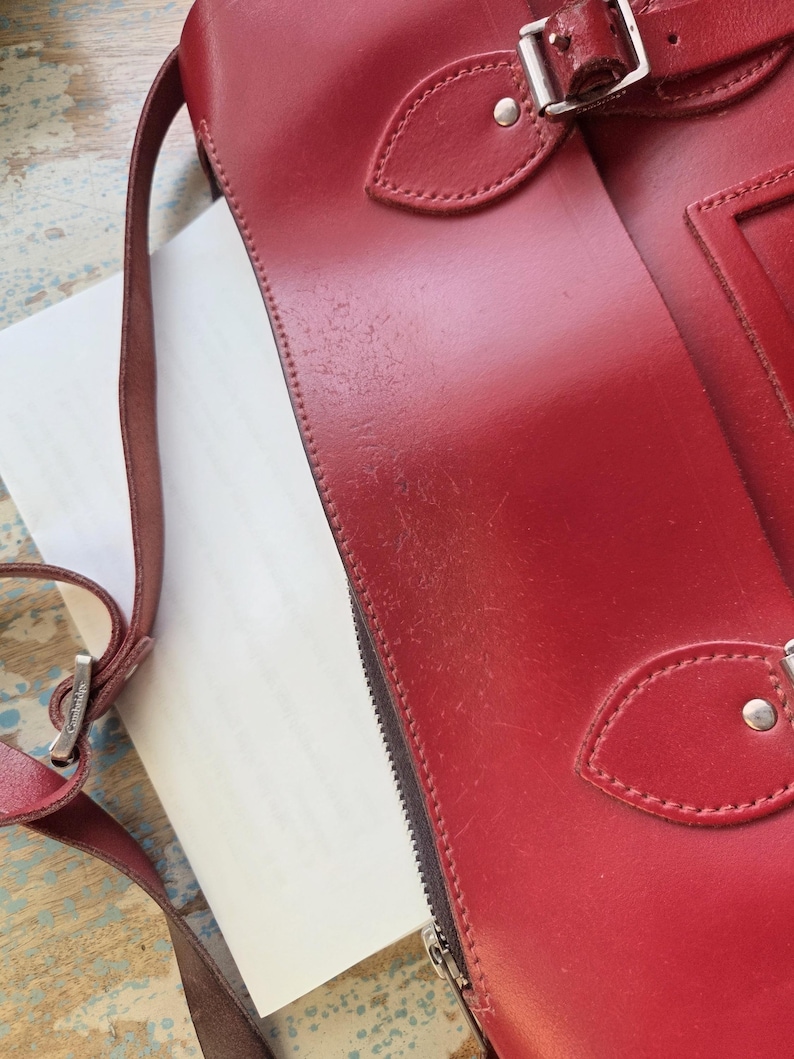 Puede incluir: Un bolso de cuero rojo con cierre de hebilla de metal plateado, una correa de hombro fina y ajustable con un conector de metal plateado grabado con la palabra Cambridge, y costuras visibles a lo largo de los bordes de la solapa.