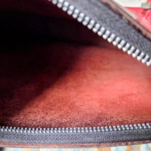 Puede incluir: El interior de un bolso con un forro texturizado de color rojo intenso. Una cremallera gris oscuro con dientes plateados recorre el borde superior de la abertura. El material parece ser cuero o ante mate y suave con grano visible.