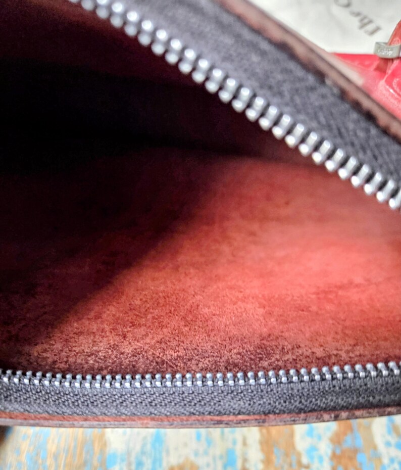Puede incluir: El interior de un bolso con un forro texturizado de color rojo intenso. Una cremallera gris oscuro con dientes plateados recorre el borde superior de la abertura. El material parece ser cuero o ante mate y suave con grano visible.