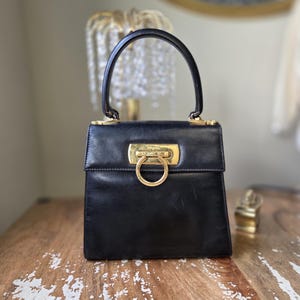 May include: A black leather handbag with a top handle and gold-tone hardware. The bag features a rectangular shape with a flap closure and a prominent gold ring detail on the front. The handle is curved and attached with gold accents.