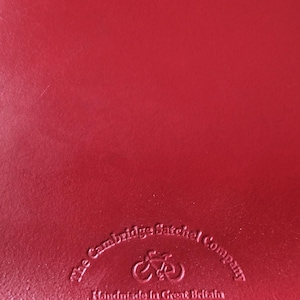 Puede incluir: Una superficie de cuero rojo liso con un logotipo en relieve que dice The Cambridge Satchel Company, con un icono de bicicleta sobre el texto y las palabras Handmade in Great Britain impresas justo debajo del gr&aacute;fico de la bicicleta.