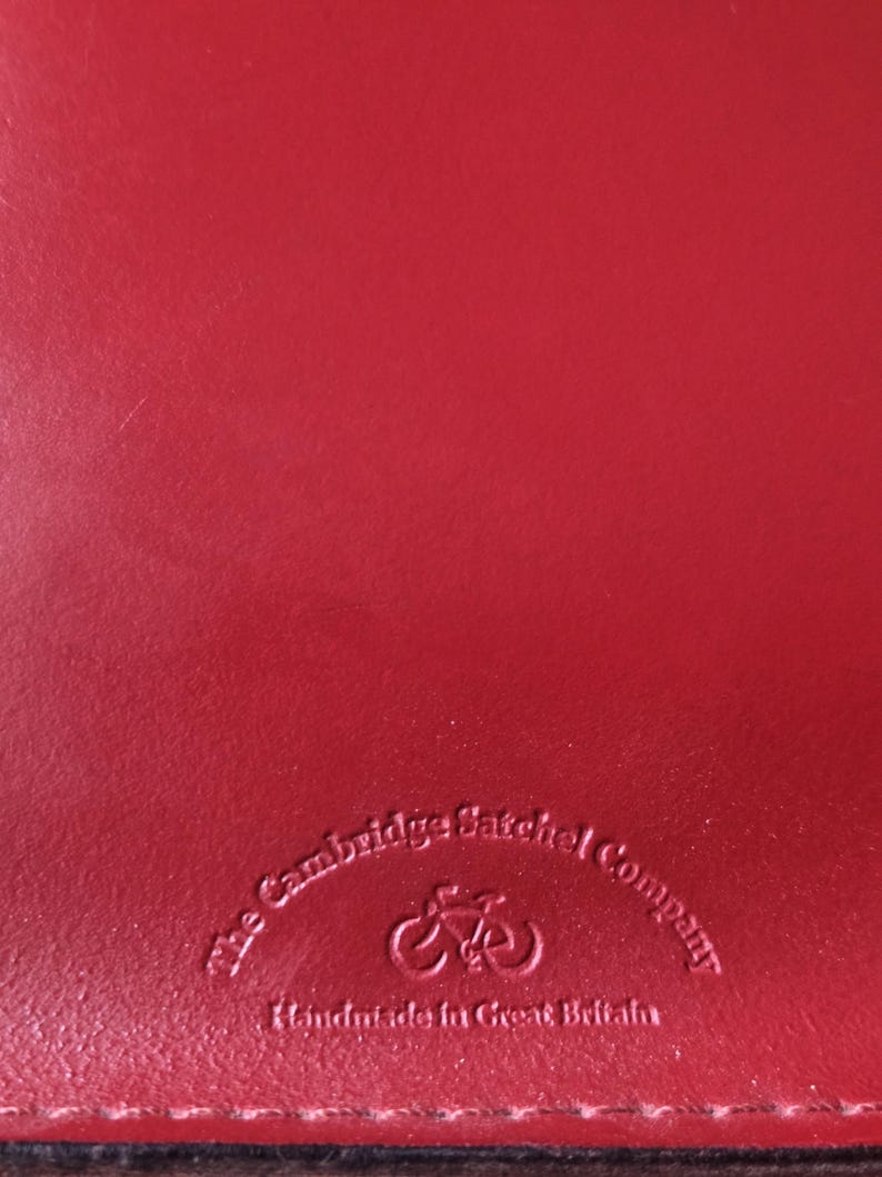 Puede incluir: Una superficie de cuero rojo liso con un logotipo en relieve que dice The Cambridge Satchel Company, con un icono de bicicleta sobre el texto y las palabras Handmade in Great Britain impresas justo debajo del gr&aacute;fico de la bicicleta.