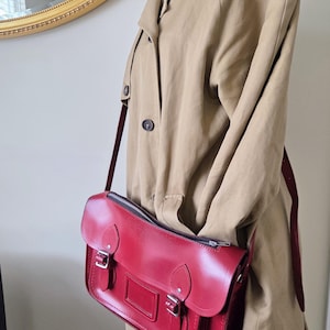 Puede incluir: Un bolso tipo cartera de cuero rojo brillante con cierre de cremallera superior, dos correas con hebilla en la parte delantera y un portaetiquetas rectangular. El bolso tiene herrajes de metal plateado y una correa larga ajustable sobre una gabardina beige.
