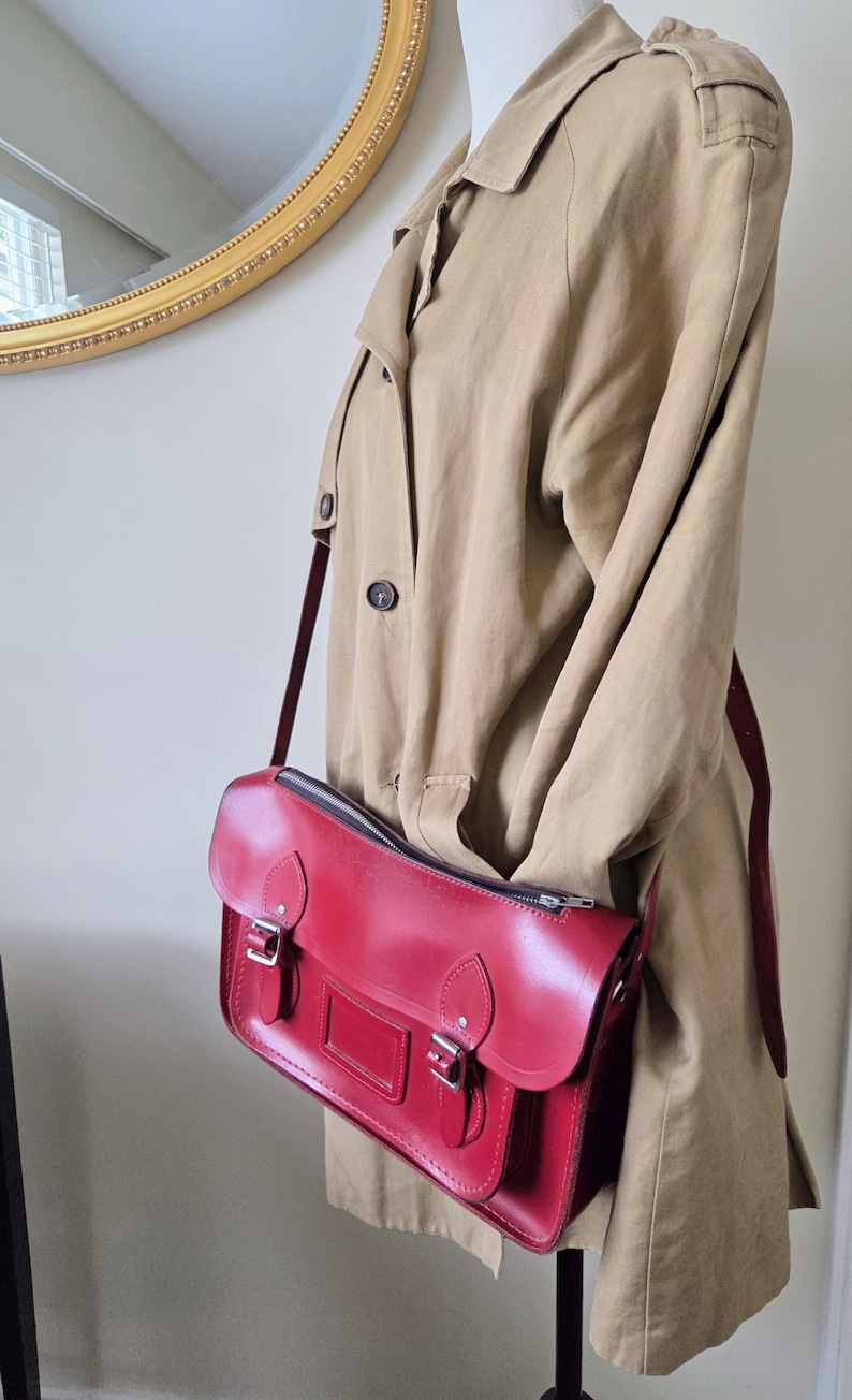 Puede incluir: Un bolso tipo cartera de cuero rojo brillante con cierre de cremallera superior, dos correas con hebilla en la parte delantera y un portaetiquetas rectangular. El bolso tiene herrajes de metal plateado y una correa larga ajustable sobre una gabardina beige.