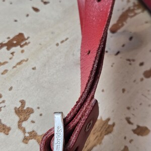 Puede incluir: Una correa de cuero rojo con costuras visibles y una hebilla de metal plateado. La hebilla tiene grabado el nombre Cambridge. La correa cuenta con varios orificios de ajuste y un remache de metal que asegura el extremo de cuero doblado.