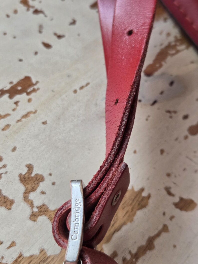 Puede incluir: Una correa de cuero rojo con costuras visibles y una hebilla de metal plateado. La hebilla tiene grabado el nombre Cambridge. La correa cuenta con varios orificios de ajuste y un remache de metal que asegura el extremo de cuero doblado.