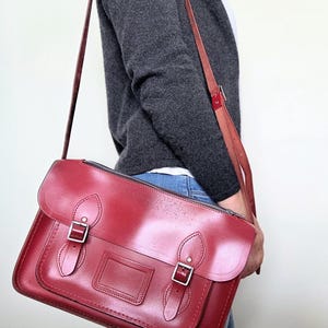 Puede incluir: Un bolso tipo cartera de cuero rojo oscuro con solapa superior, dos correas con hebilla en la parte delantera y un tarjetero rectangular central. El bolso cuenta con una correa larga ajustable con herrajes de metal plateado y cierre de cremallera.