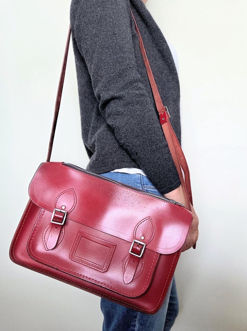 Puede incluir: Un bolso tipo cartera de cuero rojo oscuro con solapa superior, dos correas con hebilla en la parte delantera y un tarjetero rectangular central. El bolso cuenta con una correa larga ajustable con herrajes de metal plateado y cierre de cremallera.