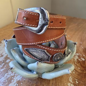 May include: A brown leather belt with a silver buckle and decorative silver accents. The belt is stitched with white thread and is coiled. The belt is resting on a light blue decorative object.