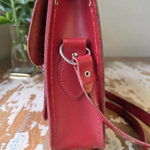 Puede incluir: Una vista lateral de un bolso tipo cartera de cuero rojo con acabado liso, solapa superior y una anilla en D de metal plateado para la correa. El bolso est&aacute; construido con costuras visibles y un remache met&aacute;lico en el conector de la correa.