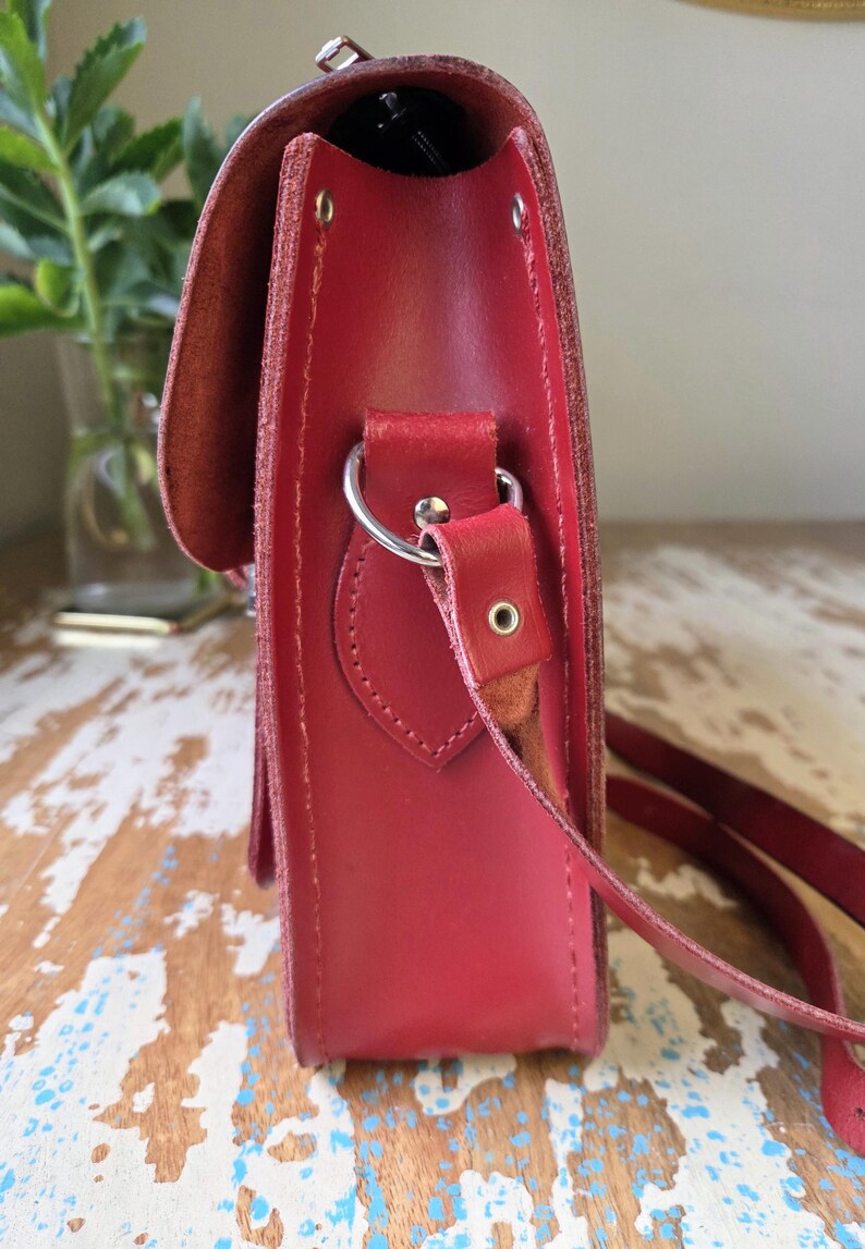 Puede incluir: Una vista lateral de un bolso tipo cartera de cuero rojo con acabado liso, solapa superior y una anilla en D de metal plateado para la correa. El bolso est&aacute; construido con costuras visibles y un remache met&aacute;lico en el conector de la correa.