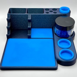 May include: A dark blue organizer with a blue mat and accents. It features compartments, a jar with a black lid, and several blue silicone rings. The organizer is designed to hold various items.