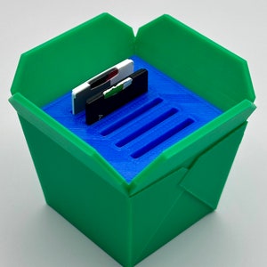 May include: A green and blue 3D printed container with a slot for holding memory cards. The container is shaped like a takeout container.