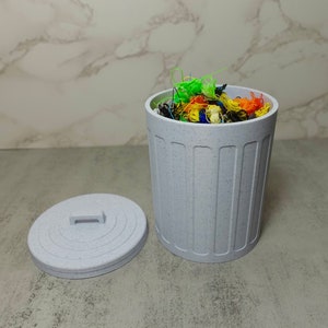 May include: A small, white, speckled trash can with a lid. The can is filled with colorful yarn scraps.