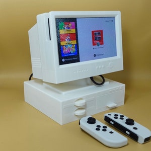May include: A white retro-style computer monitor with a screen displaying a video game interface. Two white game controllers are in front of the monitor. The monitor sits on a white base with a drawer.
