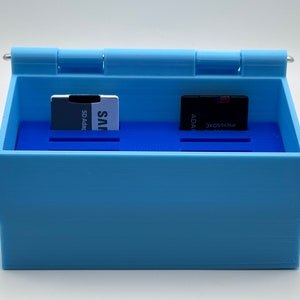 May include: A light blue plastic storage box with a hinged lid. The box has two compartments, each with a slot for a memory card. The left compartment contains a SD adapter card with the text "JVS SD Adapter" printed on it. The right compartment contains a microSDXC card with the text "MICROSDXC ADATA" printed on it.