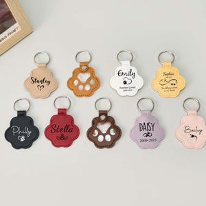 May include: A collection of paw-shaped keychains in various colors, including tan, black, red, brown, white, yellow, lavender, and pink. Each keychain features a silver ring and personalized text, such as names and dates, in a script font.