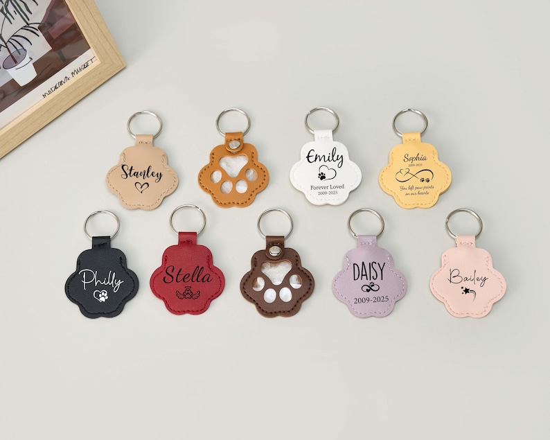 May include: A collection of paw-shaped keychains in various colors, including tan, black, red, brown, white, yellow, lavender, and pink. Each keychain features a silver ring and personalized text, such as names and dates, in a script font.
