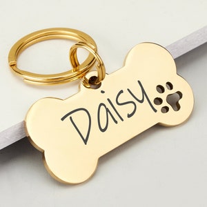 Può includere: Medaglietta per cani a forma di osso dorata con un portachiavi e il nome "Daisy" inciso su di essa. La medaglietta ha un'incisione a forma di impronta di zampa.