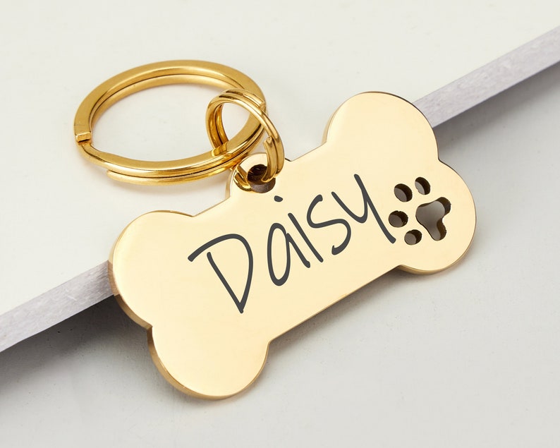May include: Gold bone-shaped dog tag with a key ring and the name "Daisy" engraved on it. The tag has a paw print cutout.