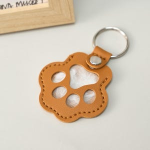 May include: A tan leather keychain in the shape of a paw print. The keychain has a silver metal ring and a small leather loop with a silver snap. The paw print has three oval cutouts and a white fluffy interior.