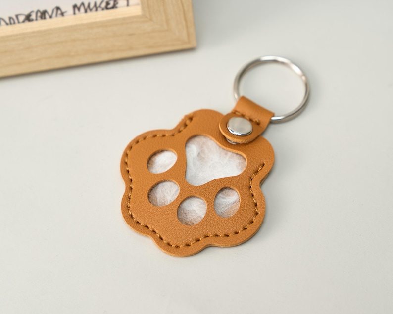 May include: A tan leather keychain in the shape of a paw print. The keychain has a silver metal ring and a small leather loop with a silver snap. The paw print has three oval cutouts and a white fluffy interior.