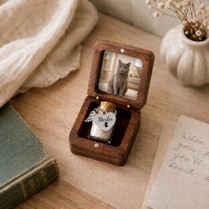 May include: A dark wood keepsake box with a hinged lid containing a photograph of a gray cat. Inside the box sits a small glass vial with a cork stopper, featuring a silver heart-shaped charm engraved with the name Merlin and a cat icon.